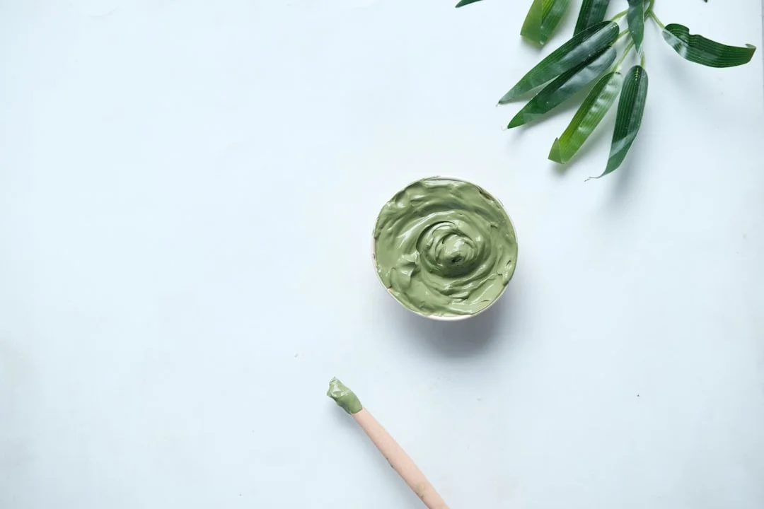 A woman relaxing with a gray-green clay mask applied to her face, a jar of clay mask and a bamboo spatula resting on a marble countertop beside her
